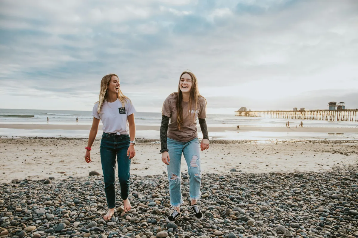 Zwei Maedchen gehen im Sommer über Kiesstrand lachend in Südengland
