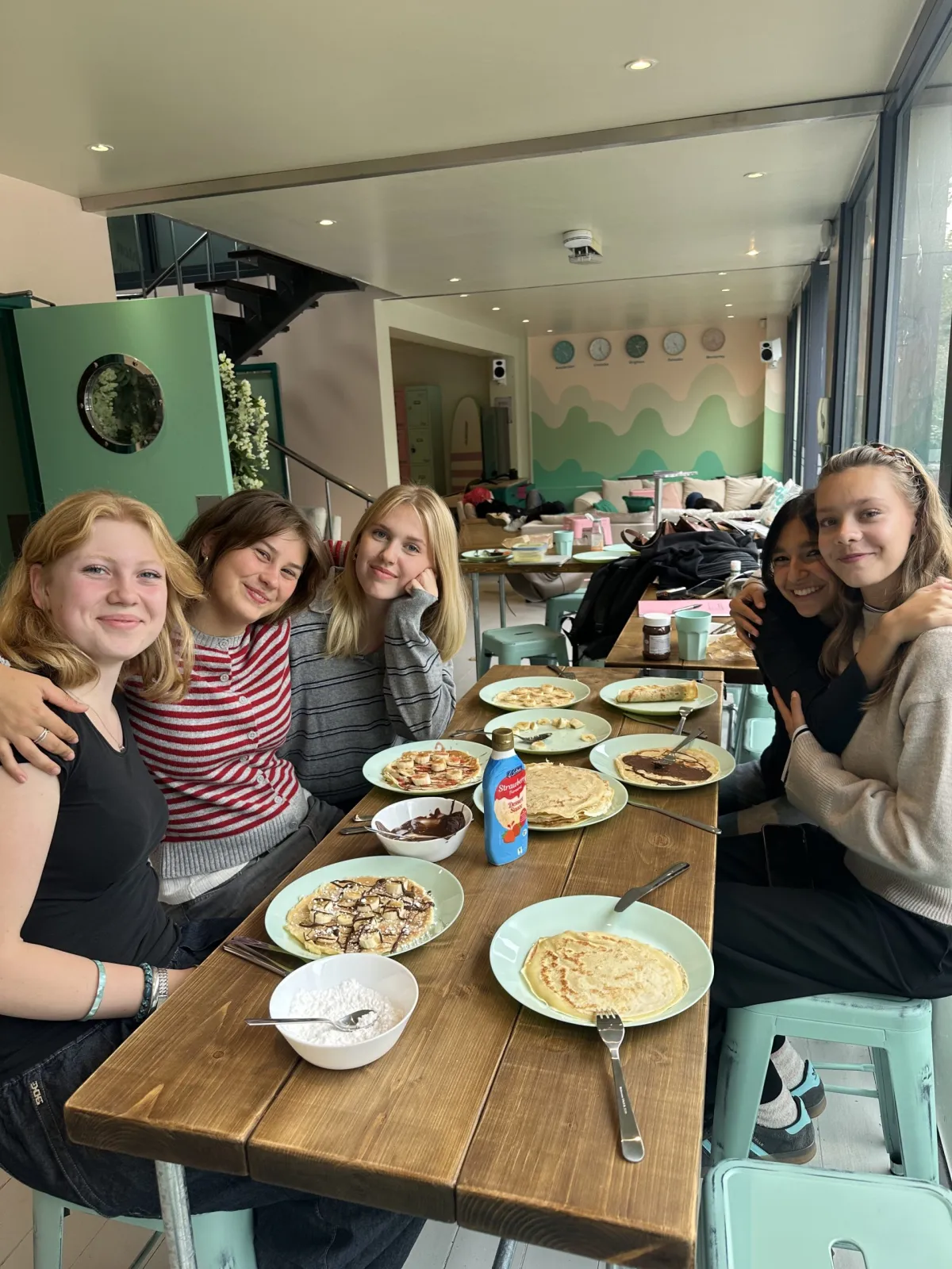 Mädchen sitzen am langen Tisch mit Pancakes auf Tellern, lachend und haltend. 