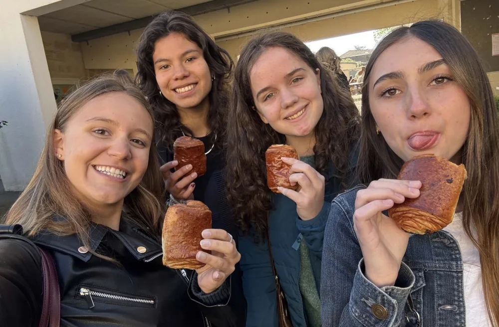 Vier junge Frauen mit Pain au Chocolat in der Hand, lachend vor einem Schulgebäude in Frankreich