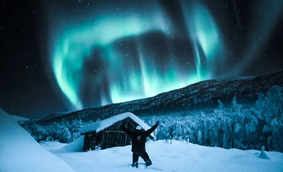Nordlichter über Hütte im Schnee mit posierendem Knabe davor