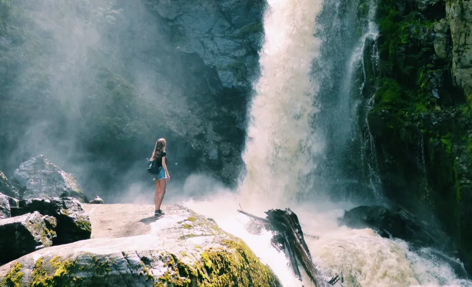 Mädchen steht unter grossem Wasserfall in Sonne mit Wasserdamp